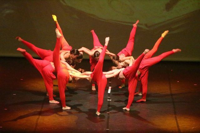 Des danseurs en justaucorps rouges forment un cercle, jambes tendues vers le haut. Scène sombre avec projecteur.