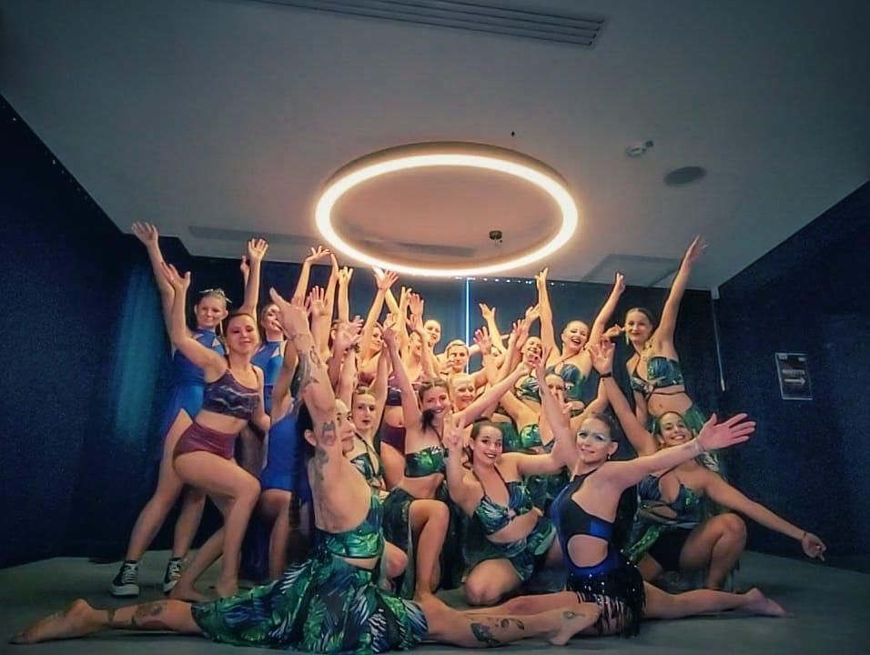 Des danseurs en costumes verts et bleus, les bras levés, dans une pièce avec un anneau lumineux au-dessus.