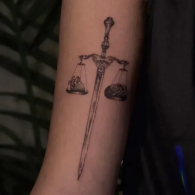 Tatuaje de una espada atravesando la balanza de la justicia en un brazo.