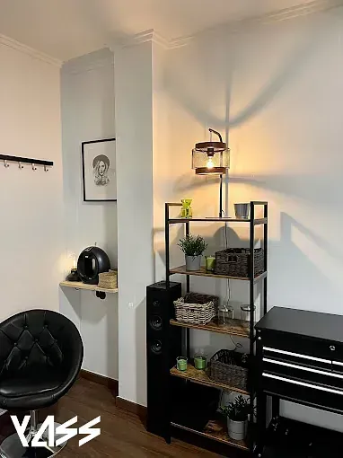 Interior acogedor con una silla negra, estantería, lámpara y obras de arte. Suelos de madera oscura y paredes en tonos neutros.