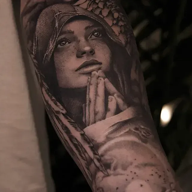 Tatuaje detallado en blanco y negro de una mujer con las manos juntas, mirando hacia arriba, encapuchada, posiblemente rezando.