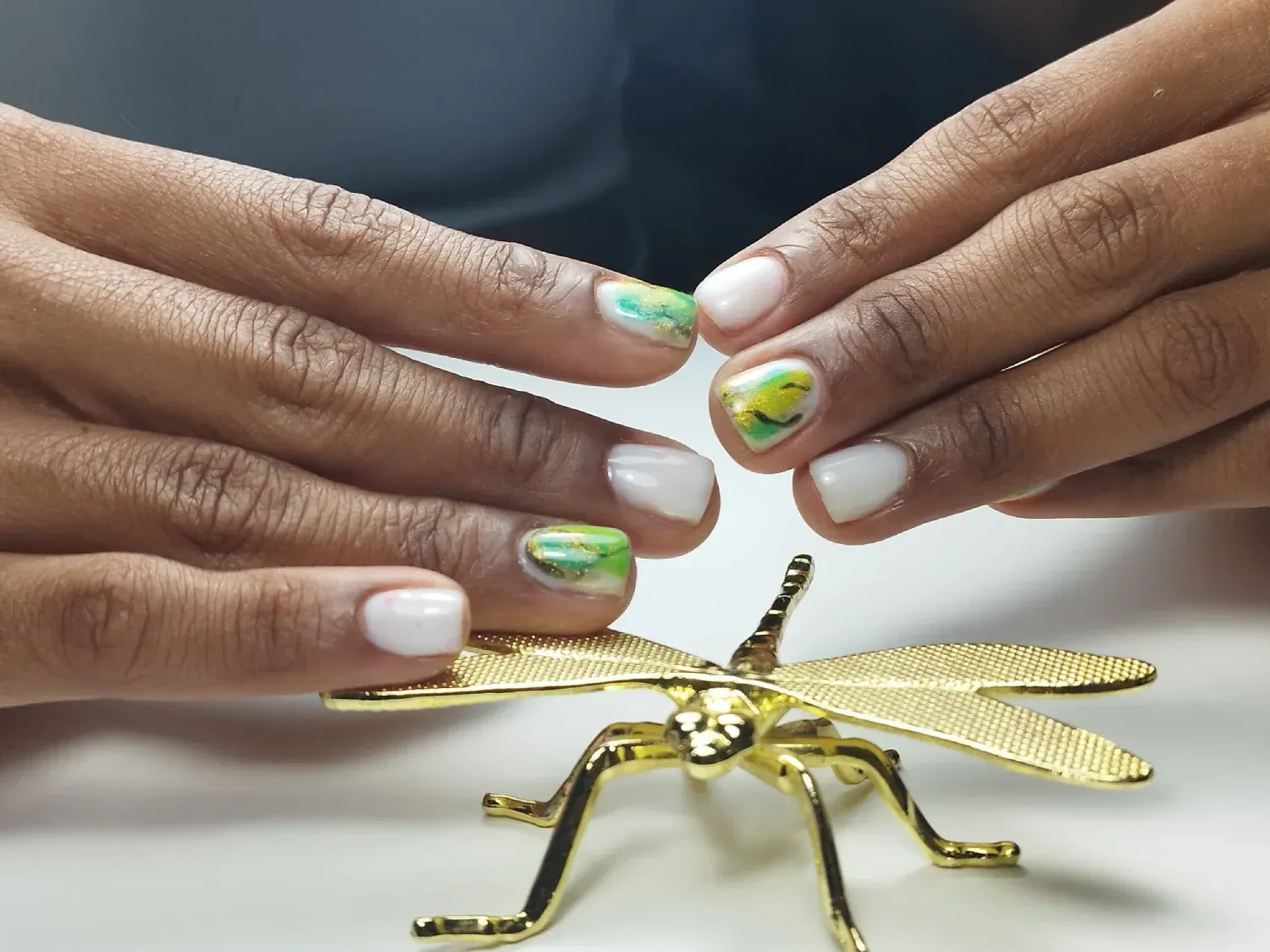 Manos con uñas decoradas en blanco y verde, sosteniendo un adorno de libélula dorada.