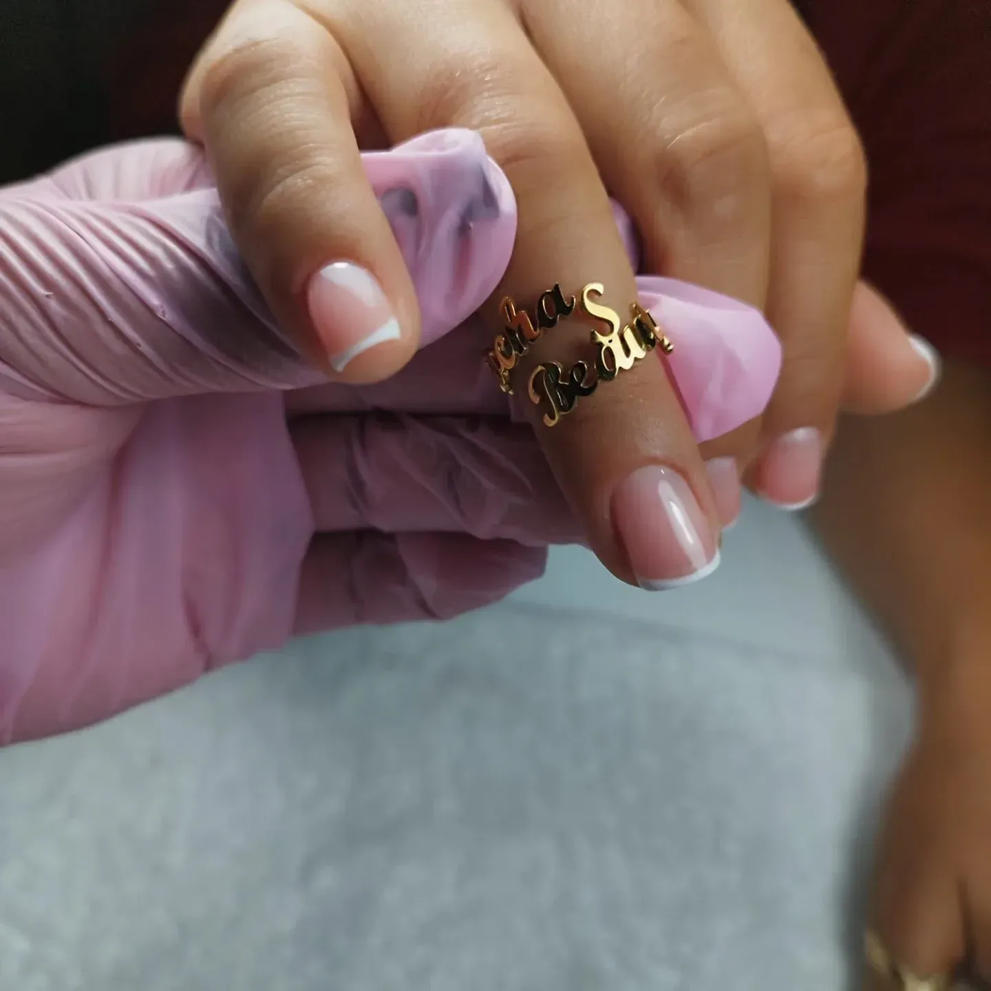 Mano que lleva un anillo de oro con uñas de punta francesa, sostenida por una mano enguantada de color rosa.