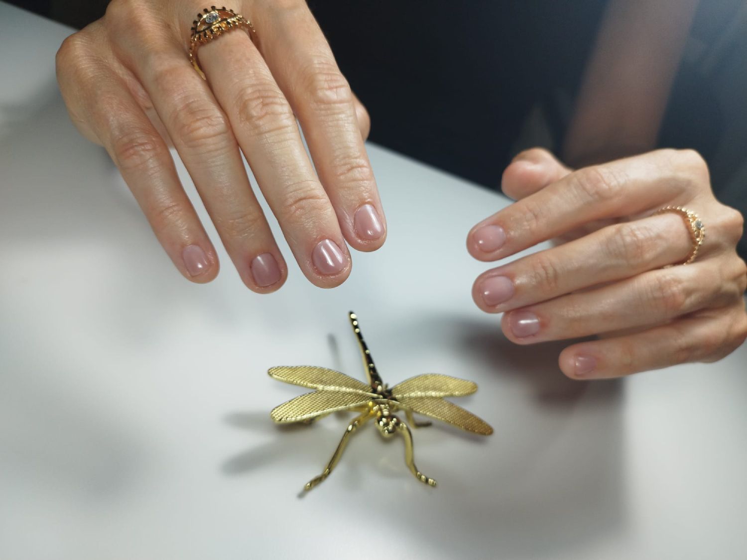 Manos con uñas esmaltadas cerca de un broche de libélula dorada sobre una superficie blanca.