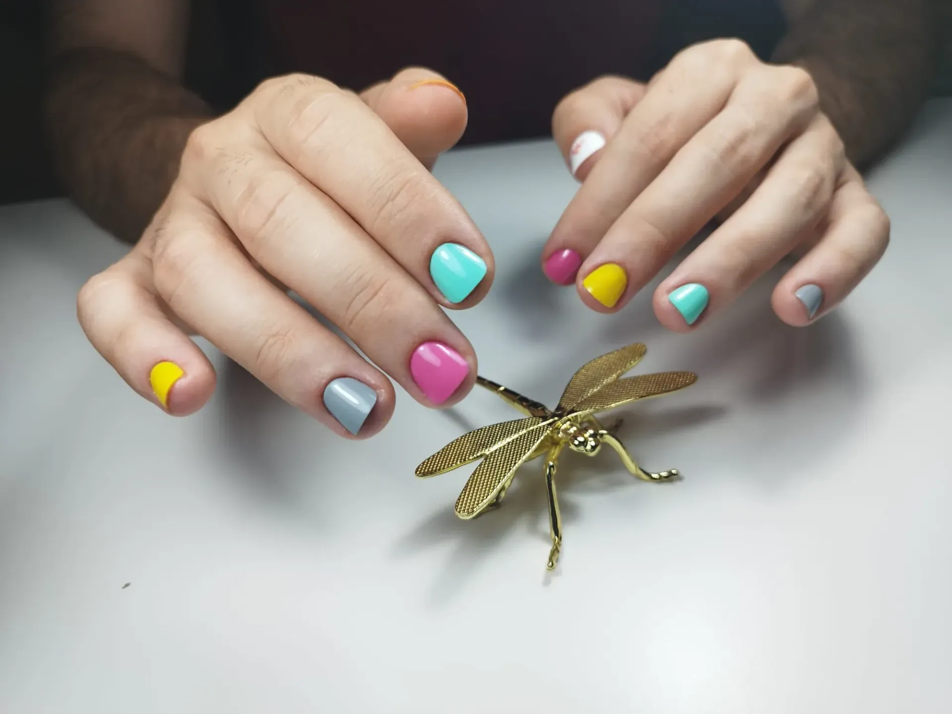 Manos con esmalte de uñas de colores, descansando cerca de una libélula dorada sobre una superficie blanca.