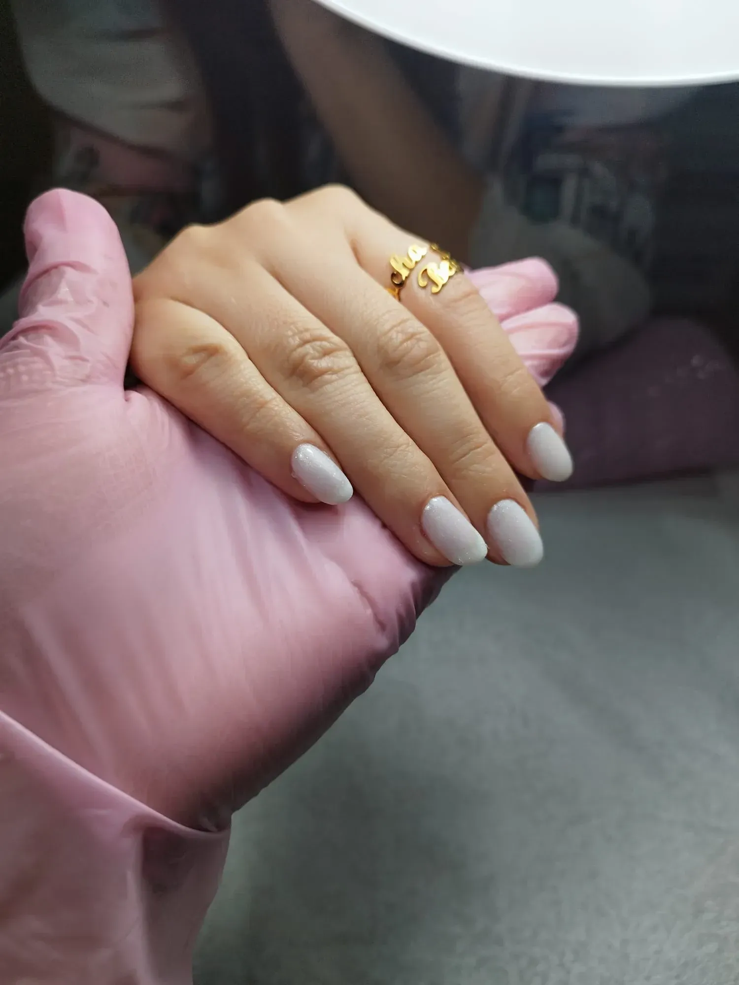 Una mano con uñas rosa claro reposa sobre una mano enguantada. Las uñas son ovaladas y lleva un anillo de oro.