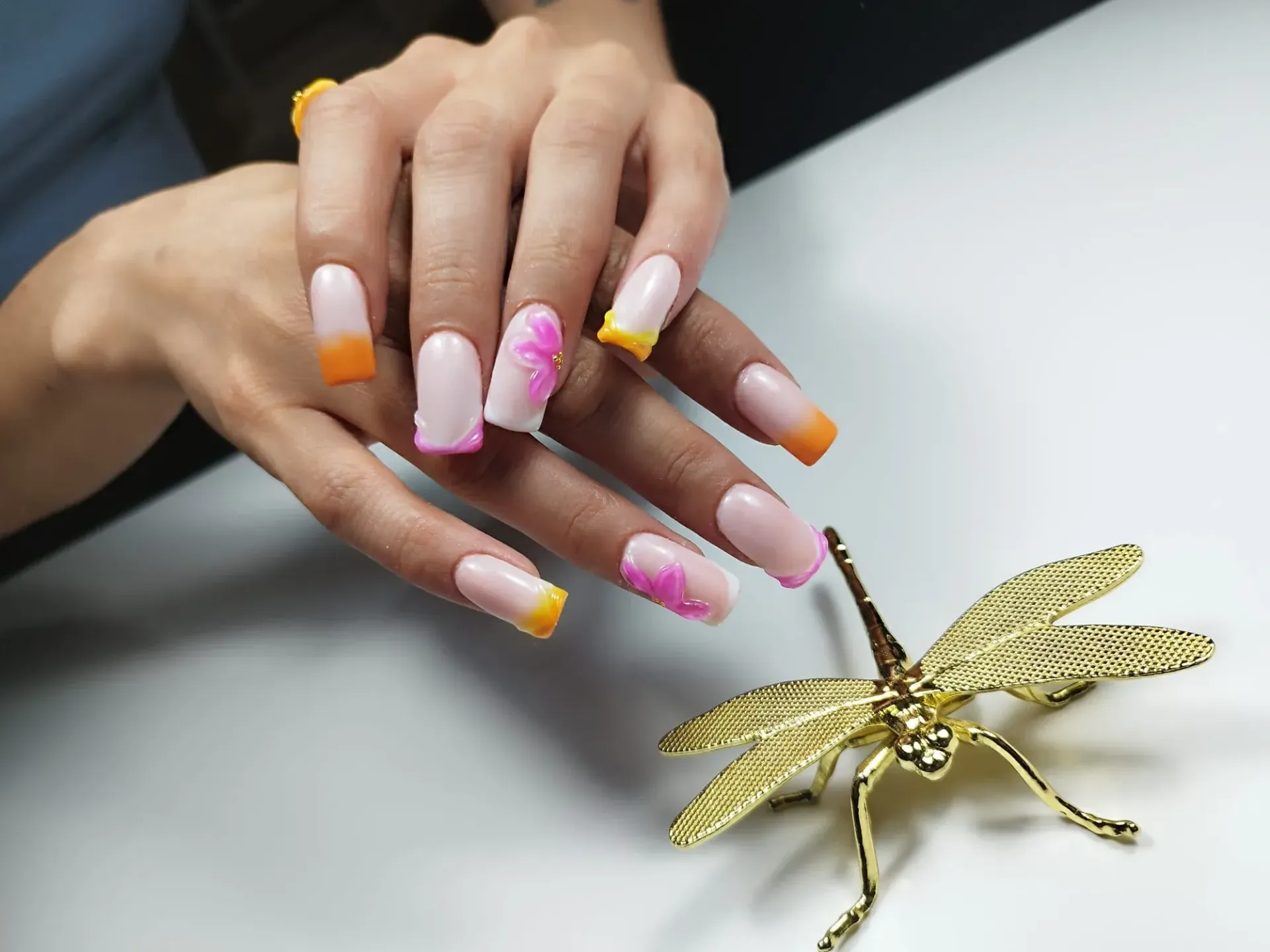 Manos con uñas acrílicas de punta cuadrada de color rosa, naranja y amarillo; adorno de libélula dorada.