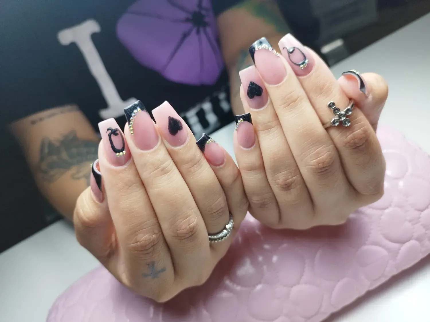 Manos con uñas acrílicas largas en forma de ataúd. Puntas negras, corazones negros y detalles plateados sobre fondo rosa.