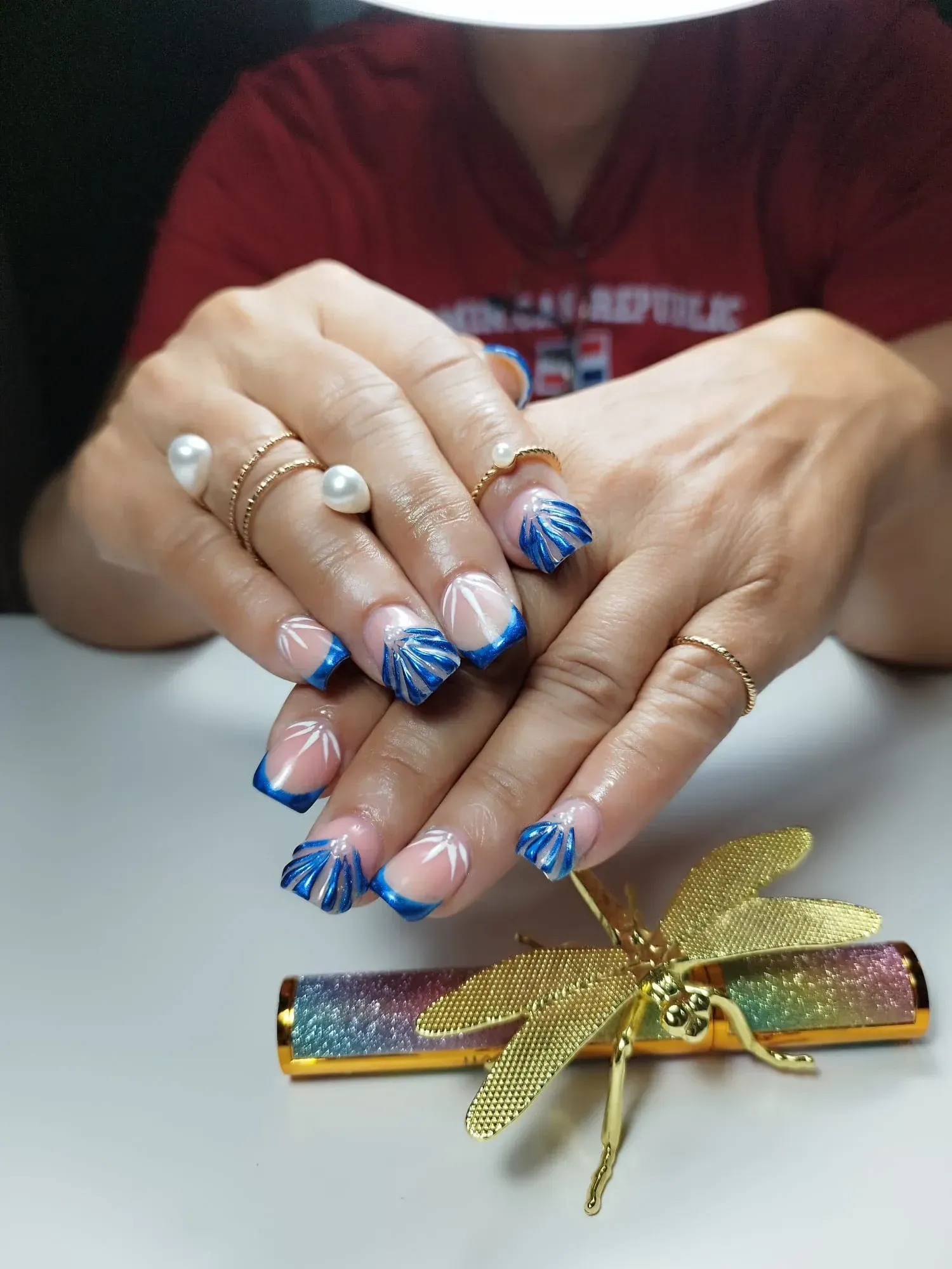 Manos con uñas decoradas estilo concha azul, con anillos dorados, apoyadas sobre una superficie
