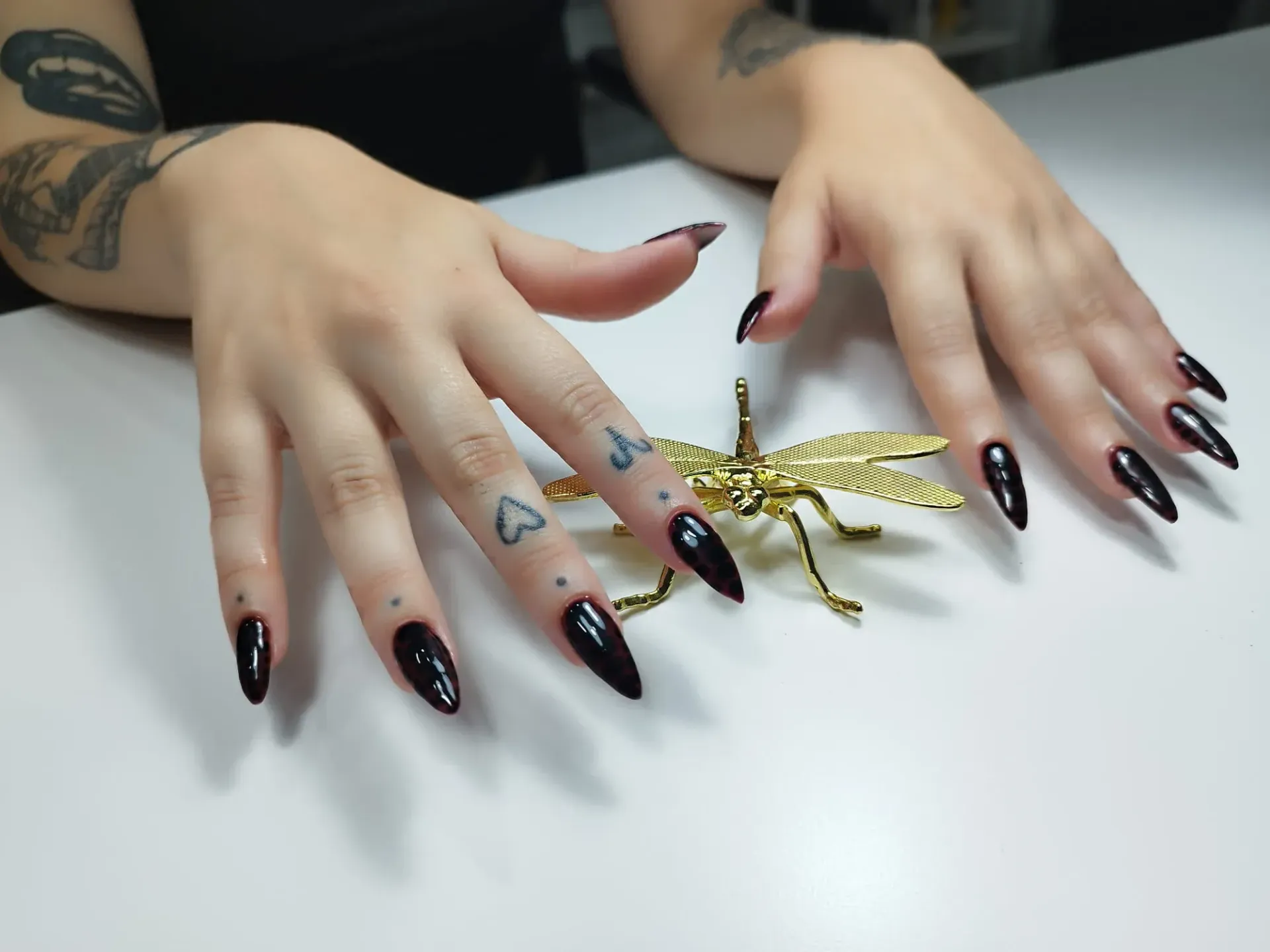 Manos con largas uñas de aguja negras, tatuajes, apoyadas sobre una superficie blanca con una figura de libélula dorada.