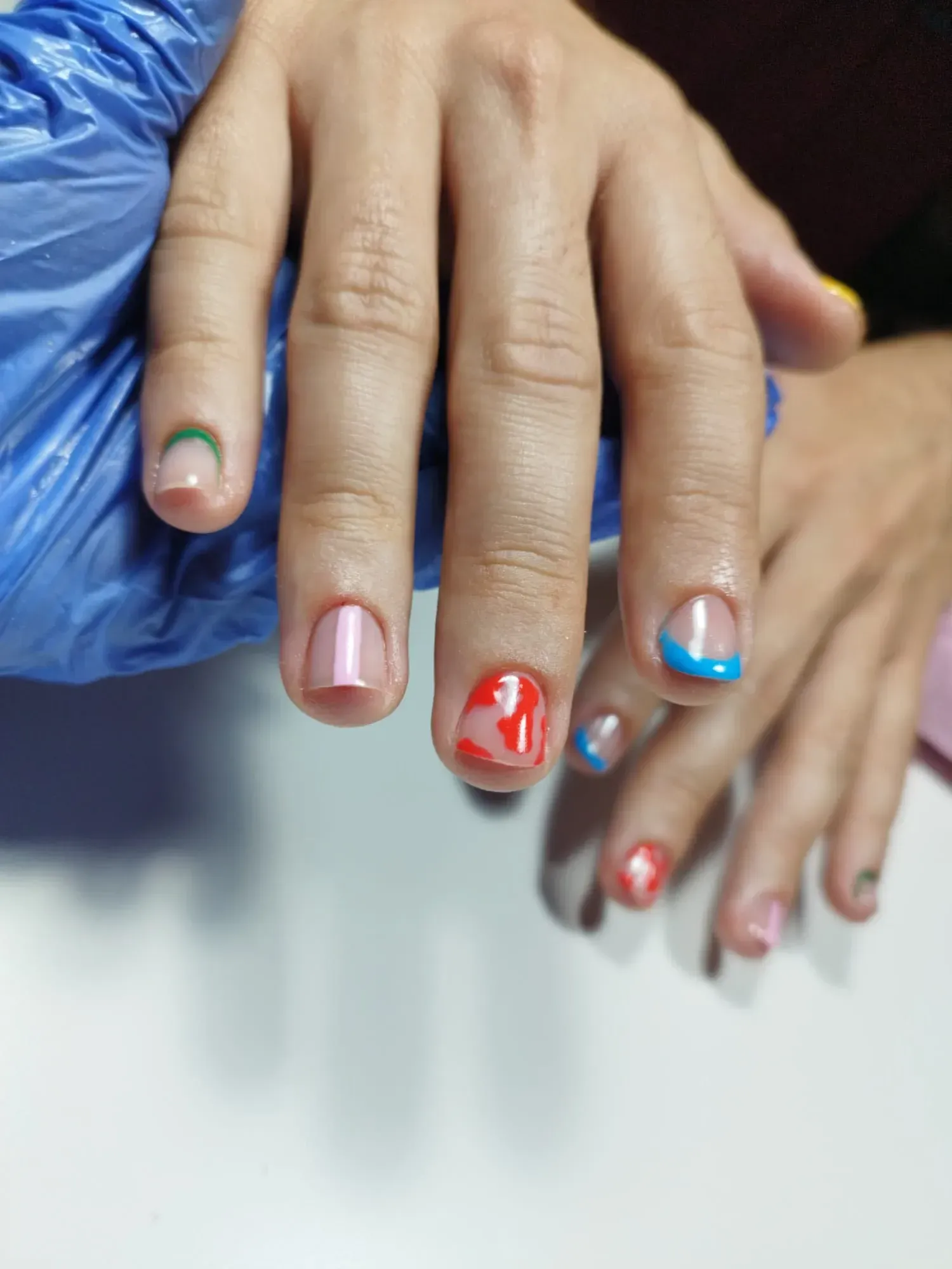 Manos con uñas cuidadas, cada una con la punta de un color diferente: verde, rosa, rojo, azul y una pequeña rosa/azul.