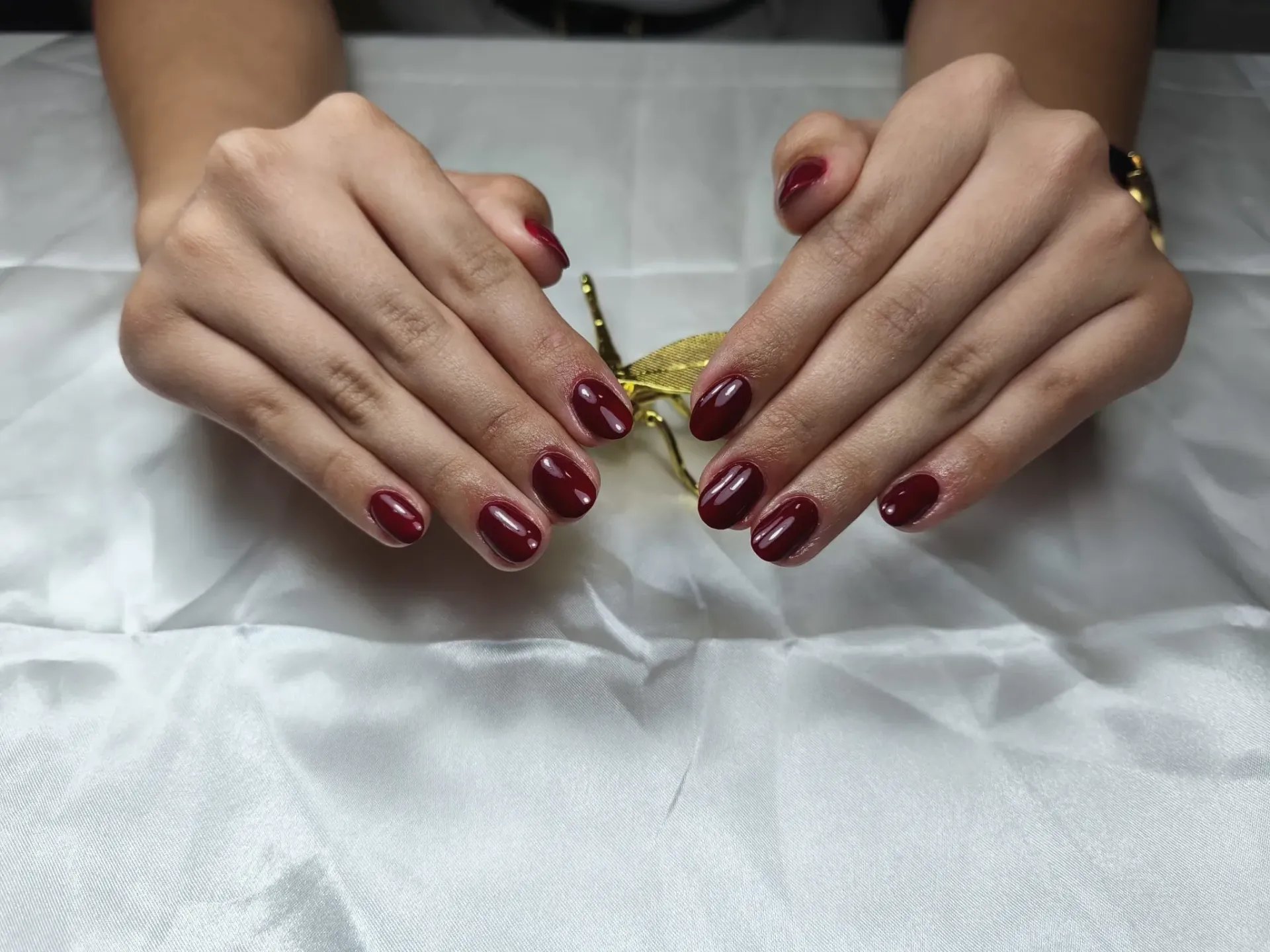 Manos con uñas brillantes de color rojo oscuro que descansan sobre tela blanca.