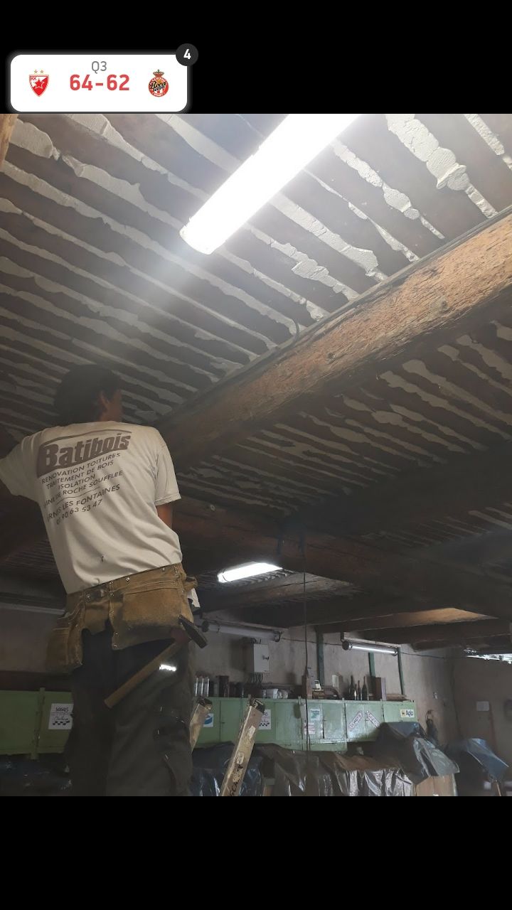 Une personne se tient debout sous un toit en tôle ondulée dans un atelier faiblement éclairé, travaillant près de lampes suspendues et d'outils.
