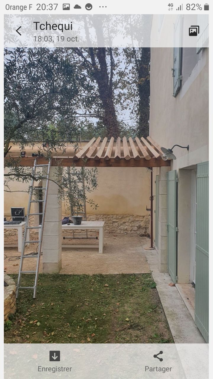 Petit patio avec une pergola en bois, une échelle, un banc et de l'herbe le long d'un mur de maison avec de grands arbres en arrière-plan.