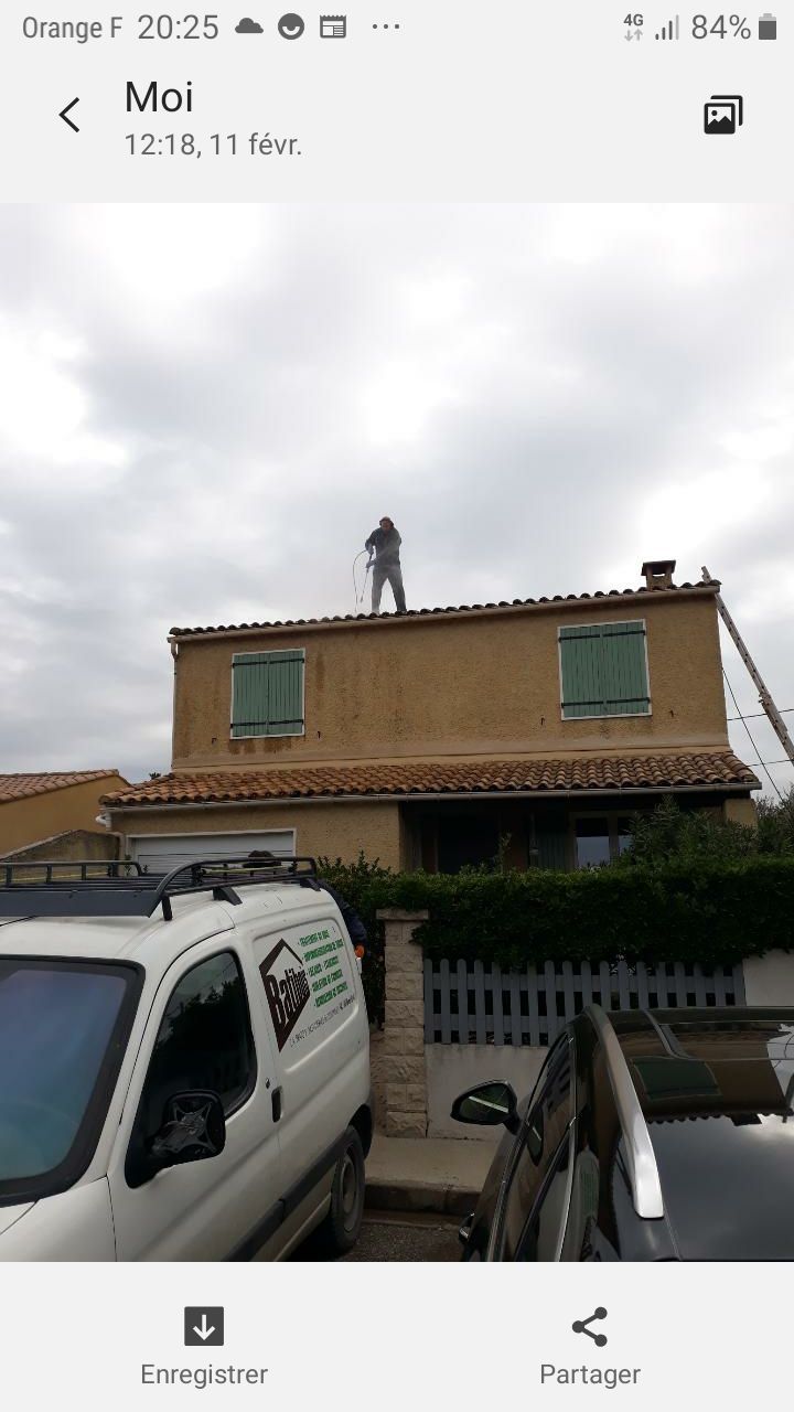 Un ouvrier se tient debout sur le toit en tuiles d'une maison à deux étages, une camionnette étant garée en contrebas sous un ciel nuageux.