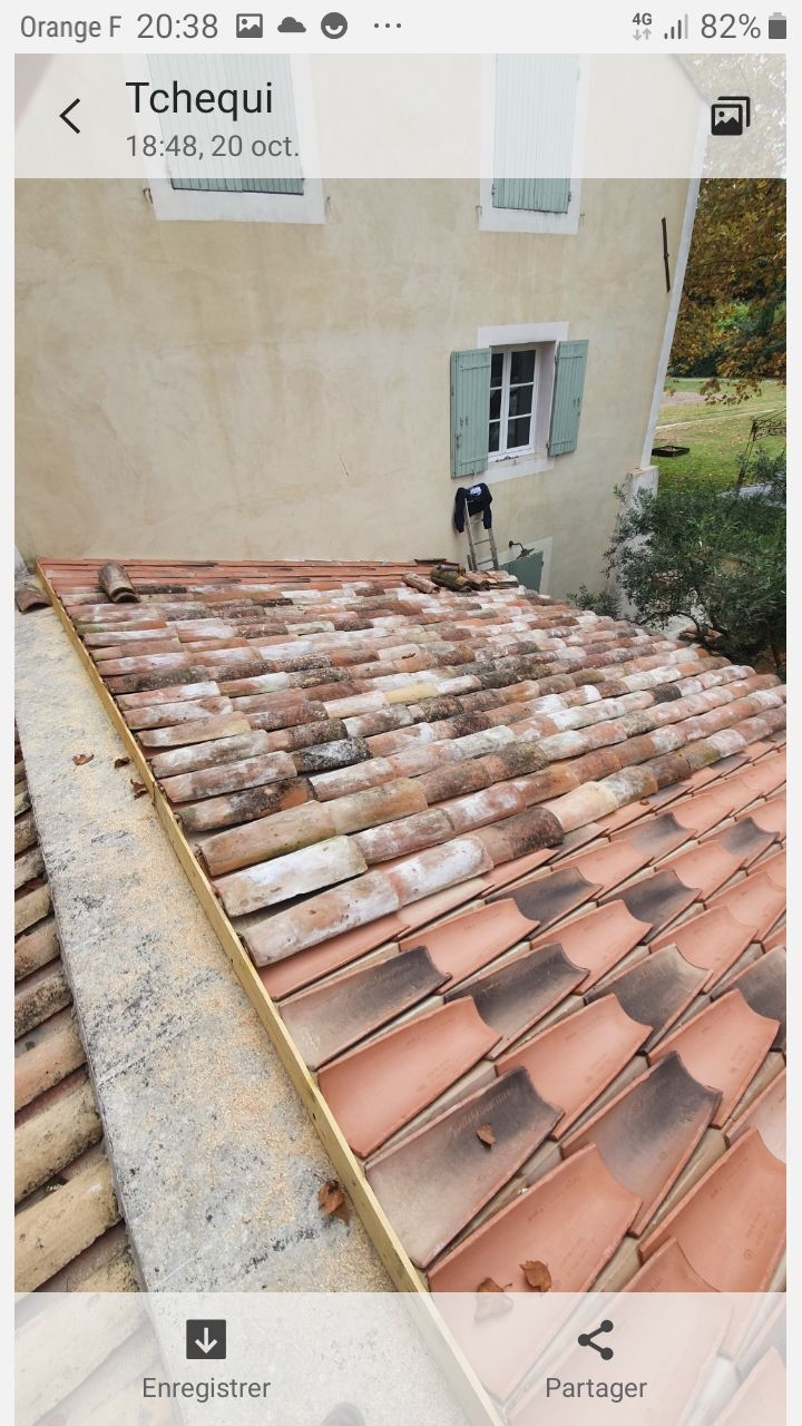 Toit en tuiles de terre cuite patinées, jouxtant un mur et une fenêtre beiges.