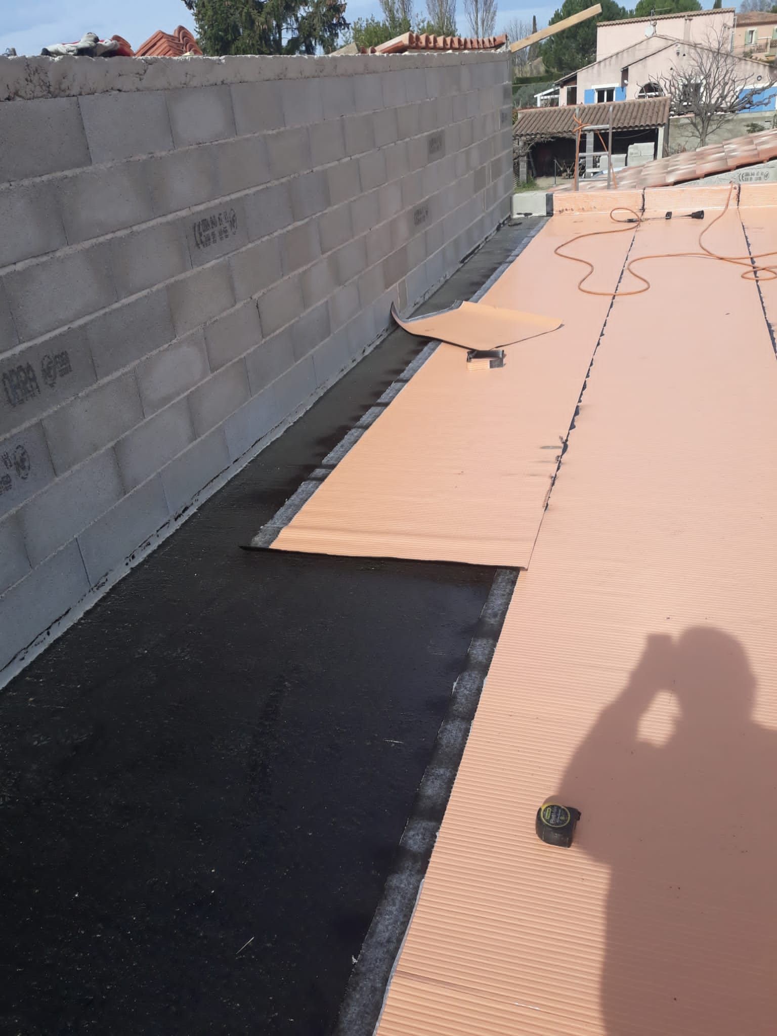 Zone de construction partiellement couverte avec mur en parpaings, sous-couche noire et tôles de toiture beiges.