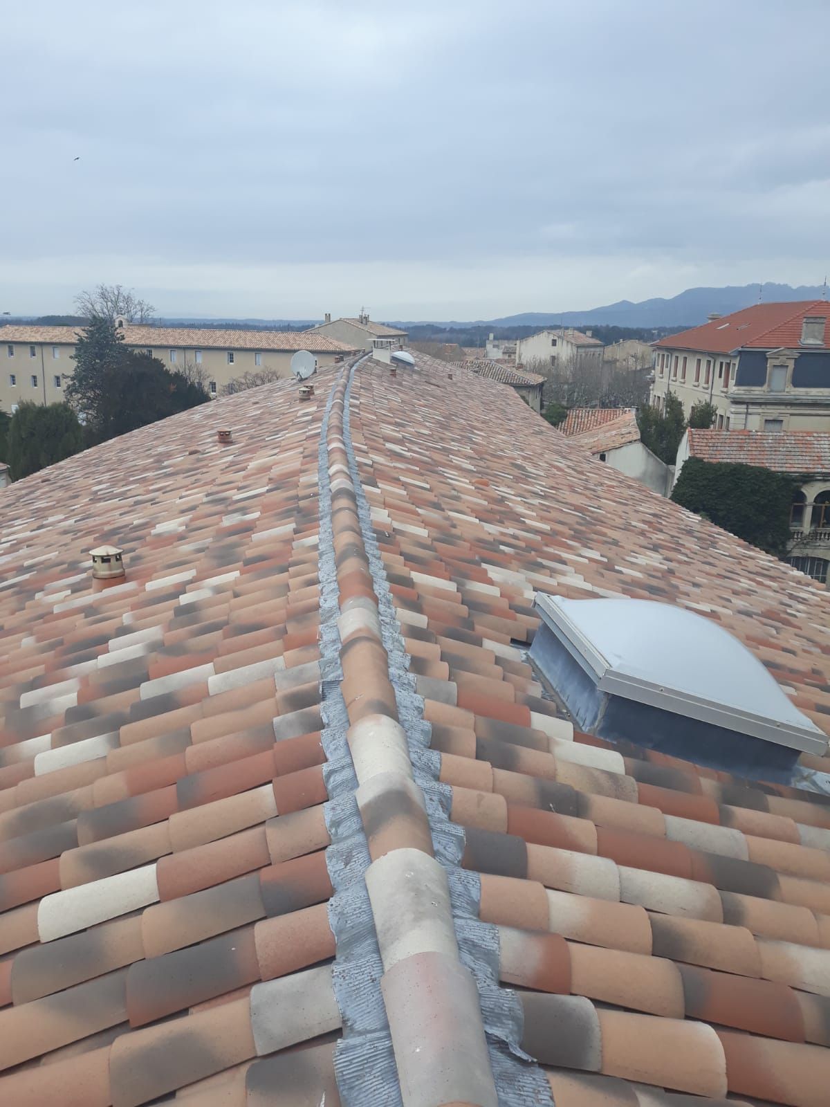 Toit-terrasse en tuiles de terre cuite avec puits de lumière, donnant sur une ville et des collines lointaines sous un ciel nuageux