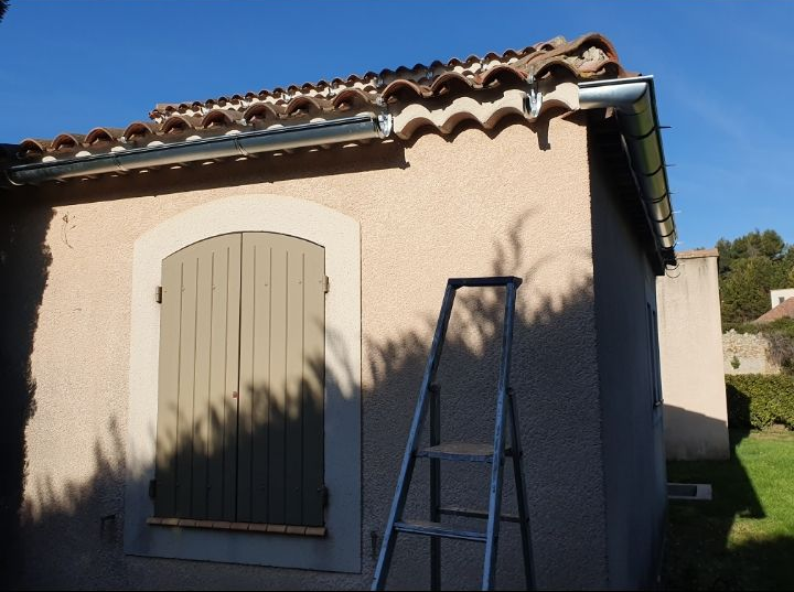 Petite maison en stuc avec toit de tuiles, porte à volets fermée et échelle appuyée contre le mur.