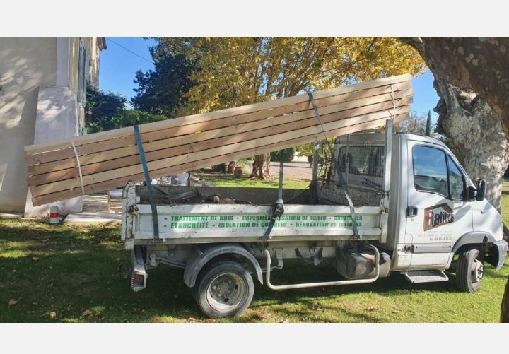 Petit camion blanc à plateau chargé de longues planches de bois dans une cour ensoleillée