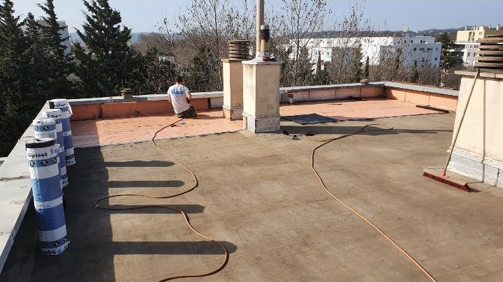 Terrasse sur le toit avec sol en béton, barils bleus le long de la gauche et un ouvrier près du mur du fond.