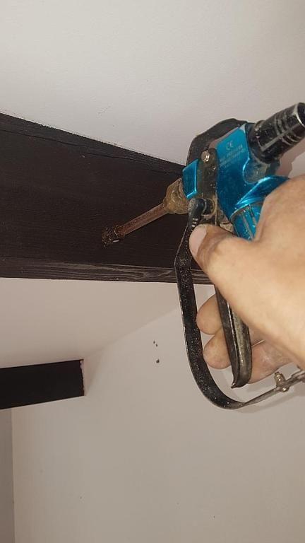 Une main utilise une perceuse bleue pour percer une poutre en bois sombre près d'un mur blanc.