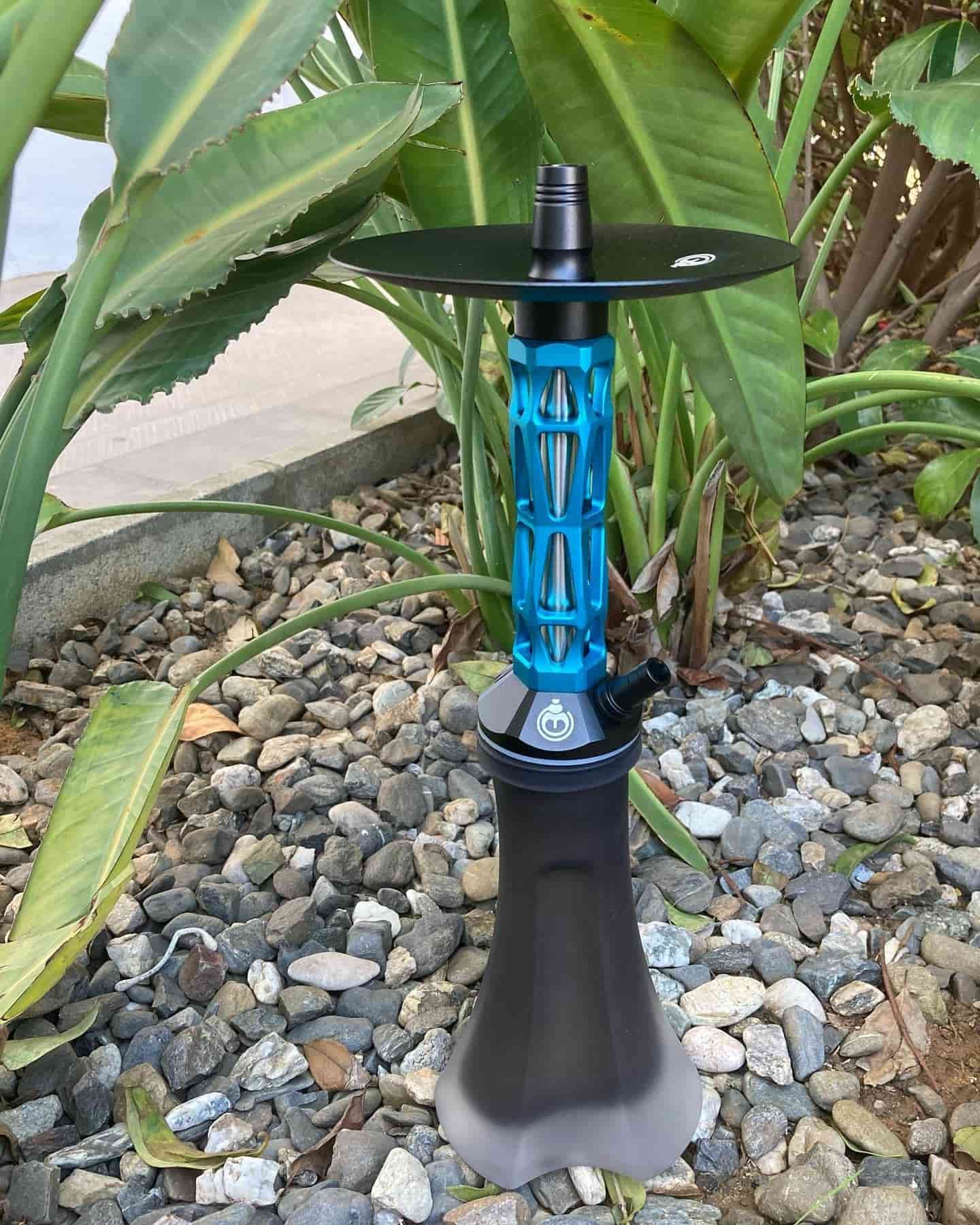 Cachimba azul y negra con bandeja grande, colocada sobre rocas junto a plantas.