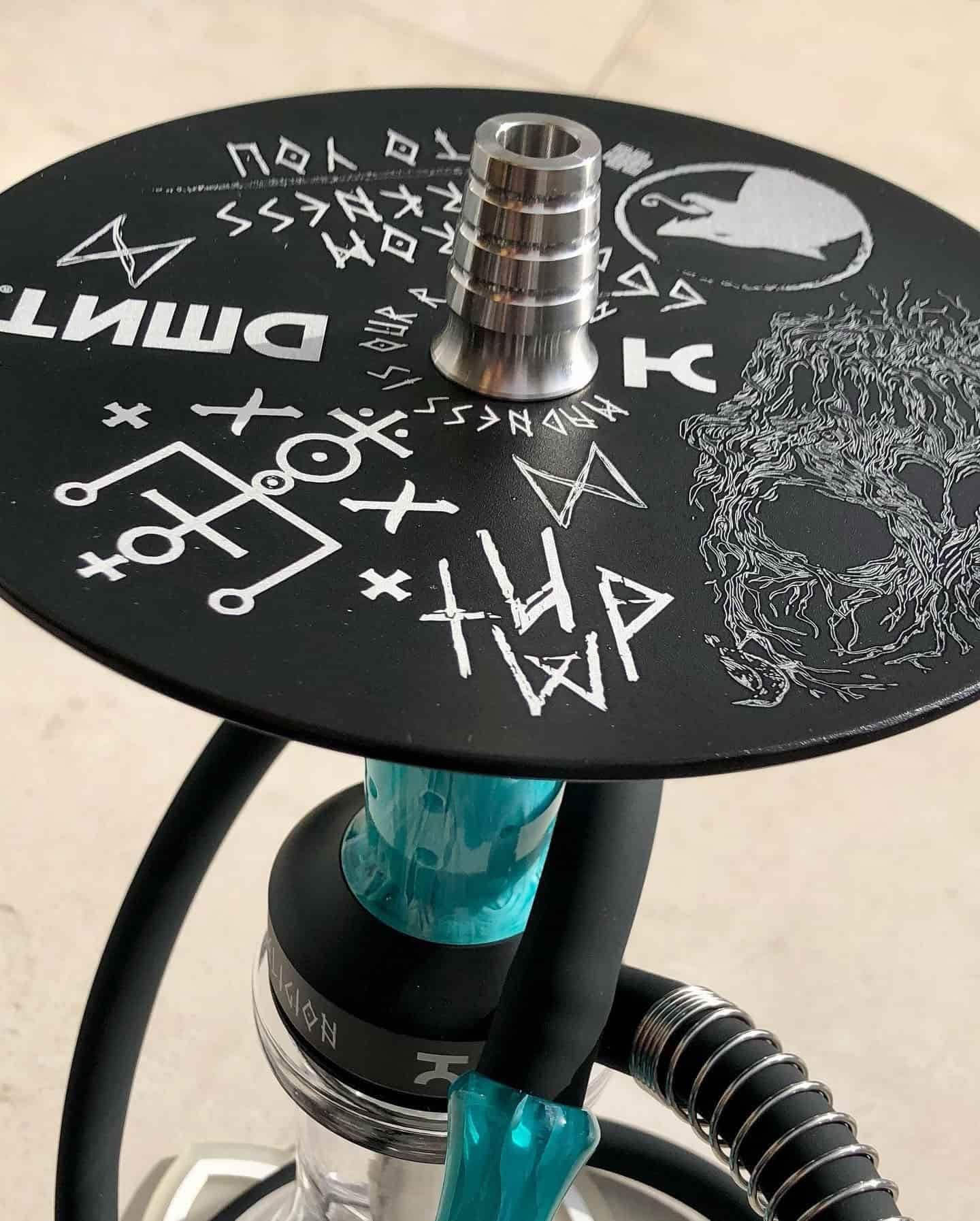 Cachimba negra con detalles en verde azulado y tapa de metal plateado. Marcas decorativas y texto en la parte superior.