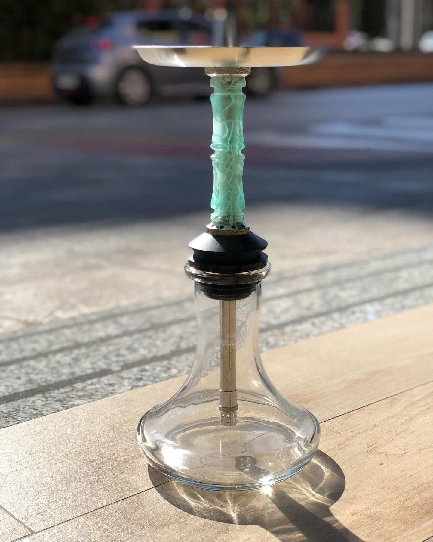 Cachimba de cristal transparente con tallo verde y bandeja de metal, colocada sobre una superficie de madera con un fondo exterior borroso.
