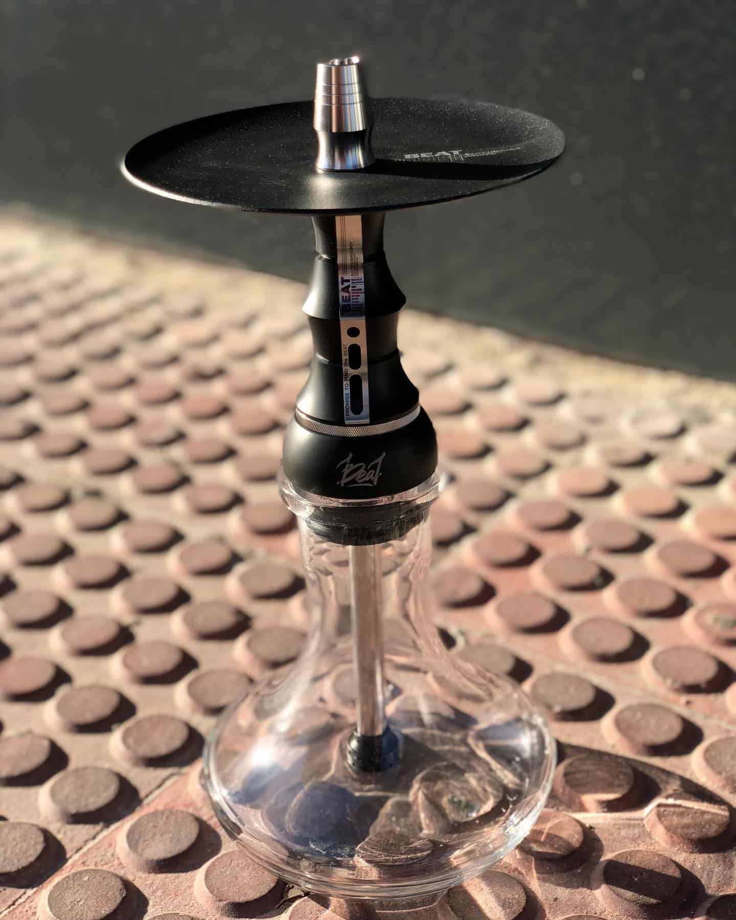 Cachimba negra y plateada con base de cristal, colocada sobre una superficie de metal texturizado.