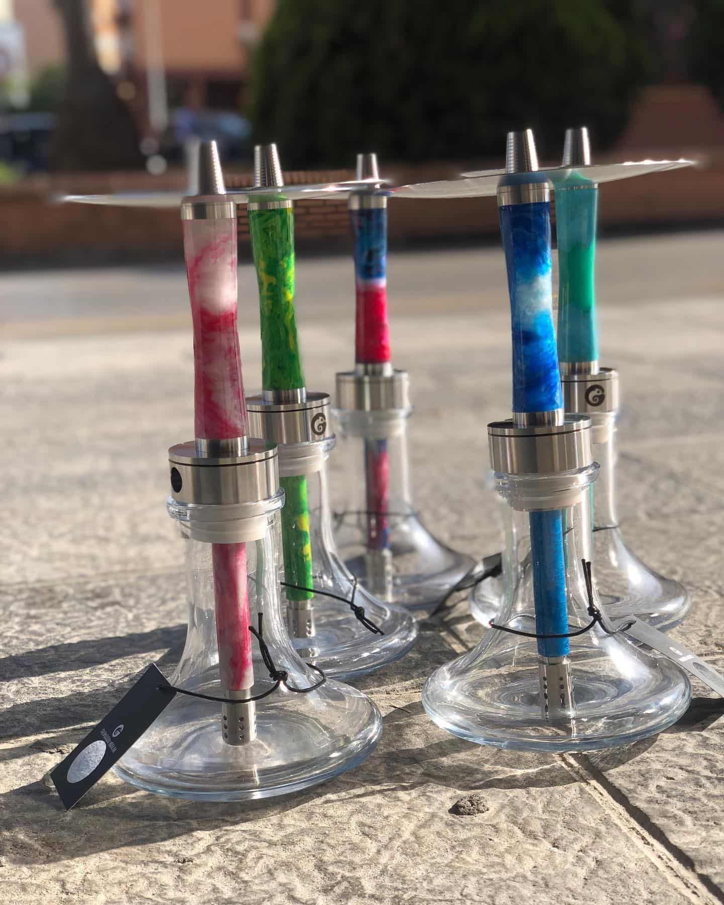 Seis coloridas cachimbas expuestas al aire libre sobre una superficie de piedra. Cada una tiene una base de vidrio transparente, detalles plateados y un vibrante tubo jaspeado.