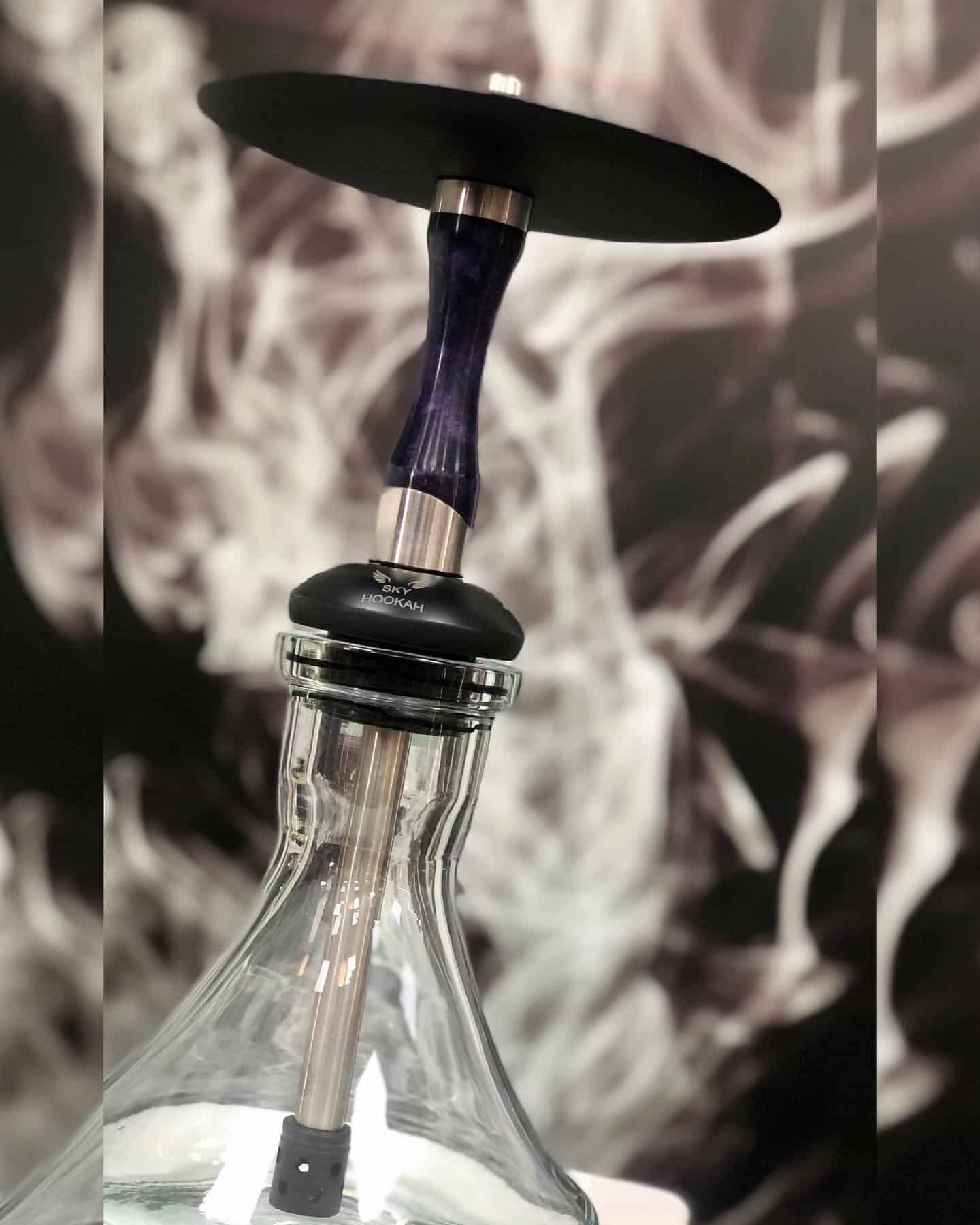 Cachimba de cristal transparente con tallo morado, bandeja negra y fondo ahumado.
