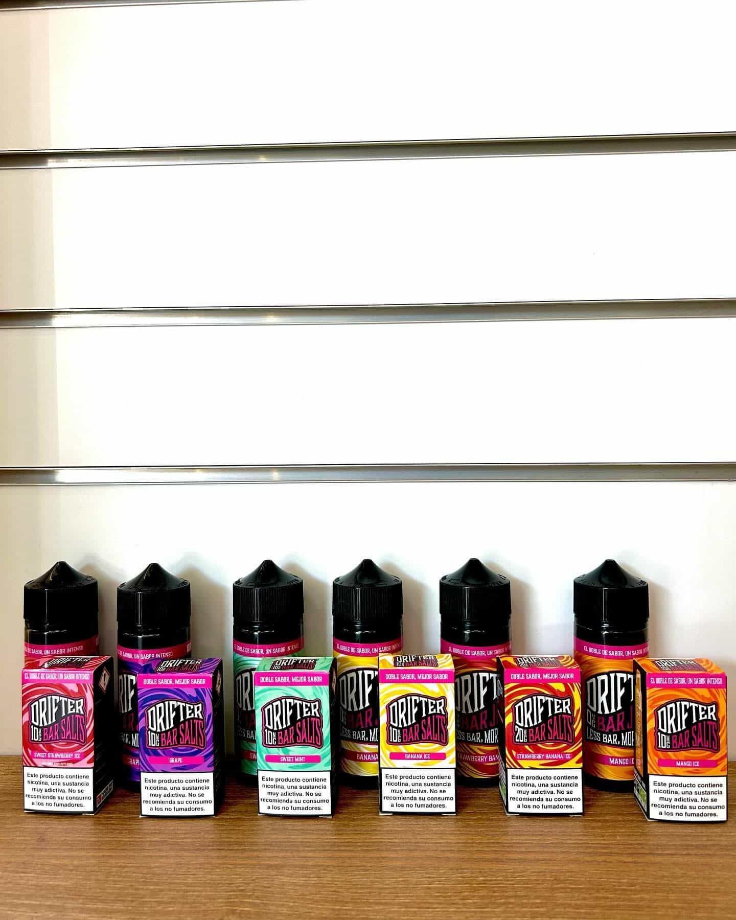 Botellas y cajas de líquido para vapear sobre una superficie de madera, contra la pared. Las botellas son mayoritariamente rosas y naranjas.