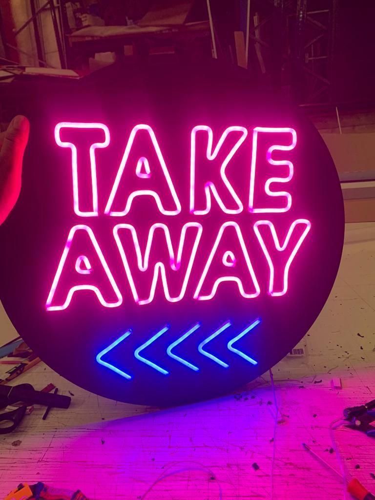 Letrero de neón: “TAKE AWAY” en color rosa, flechas azules, fondo negro circular.