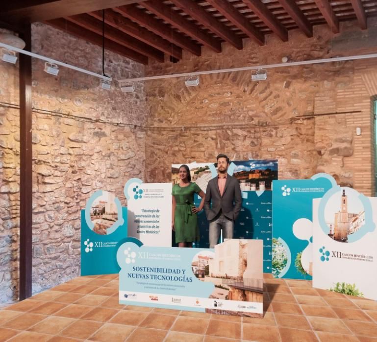 Dos personas permanecen de pie junto a un cartel de evento en una habitación con paredes de ladrillo.