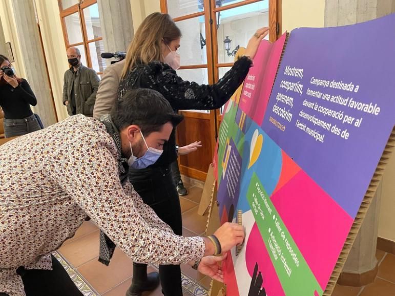 Dos personas colocan pegatinas en un colorido cartel en un interior. Una lleva una camisa floreada, todas llevan mascarillas.