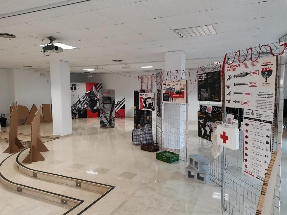 Sala de exposiciones con carteles, artefactos y estructuras de cartón. Paredes blancas, iluminación brillante.
