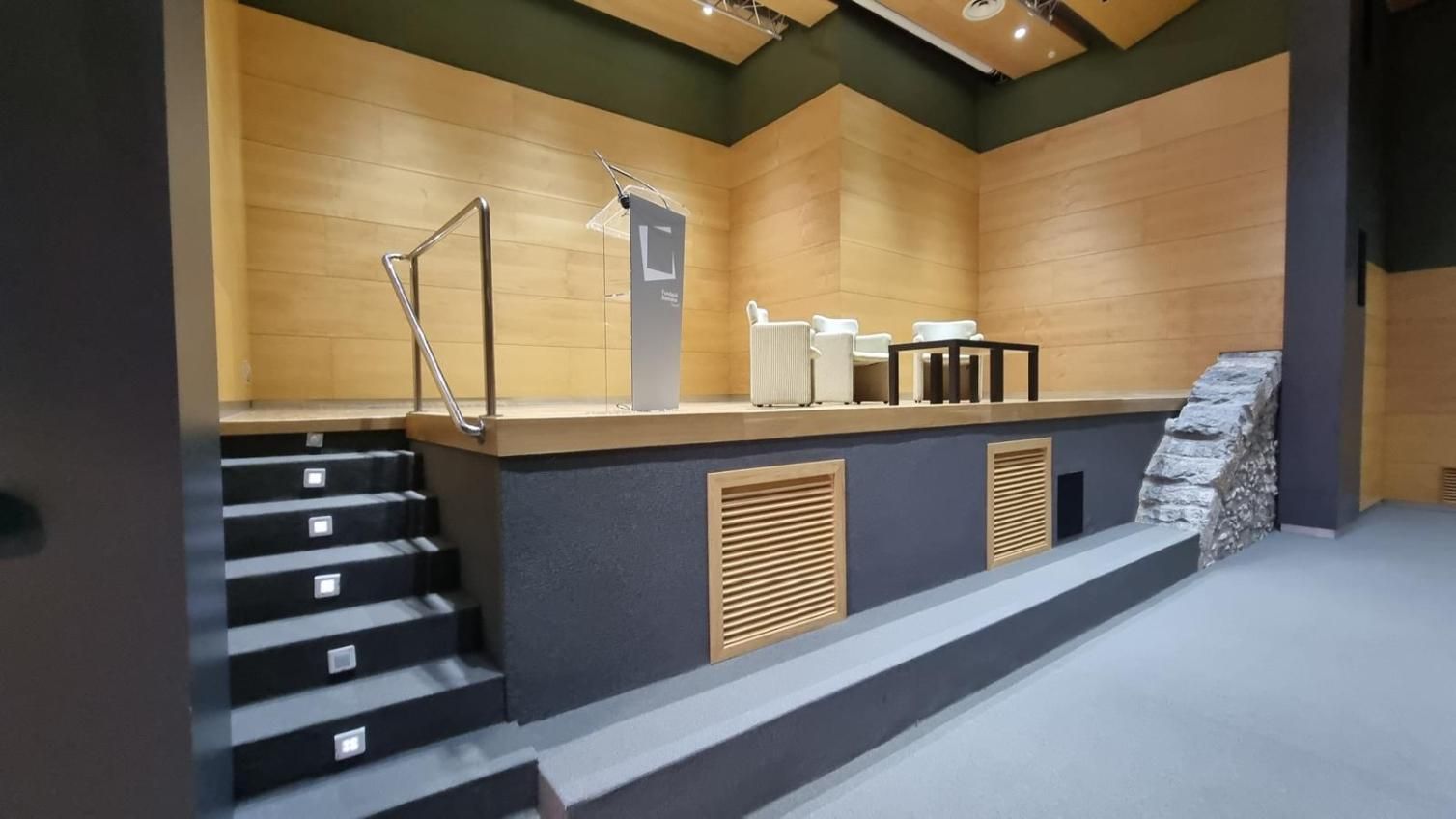 Un escenario con podio y asientos frente a una pared revestida de madera; unos escalones conducen hacia arriba.