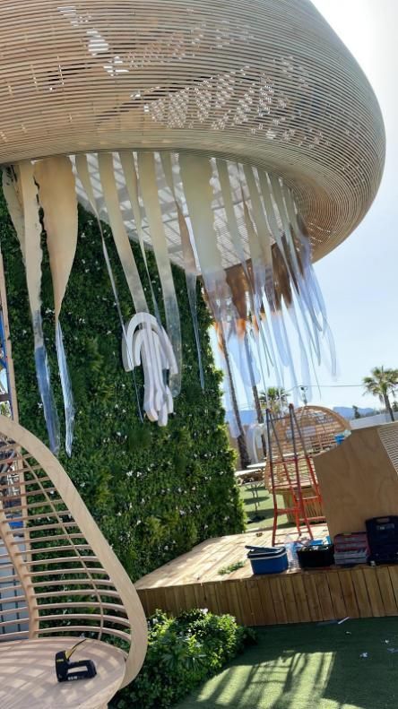 Estructura tejida con cintas fluidas sobre una pared verde, cerca de una zona de césped con muebles de exterior.