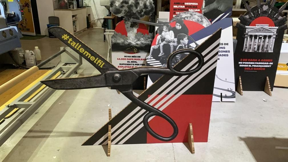 Tijeras gigantes frente a una exhibición política. Gráficos en blanco, negro y rojo. Texto y formas.