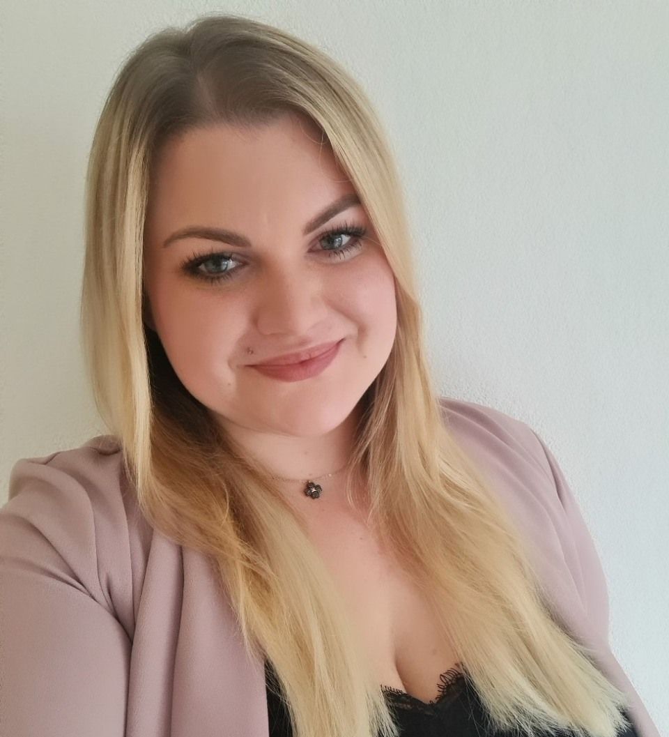 Frau mit blonden Haaren, die einen rosa Blazer trägt und lächelt.