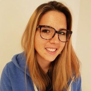 Frau mit Brille lächelt, sie trägt einen blauen Kapuzenpullover vor hellem Hintergrund.