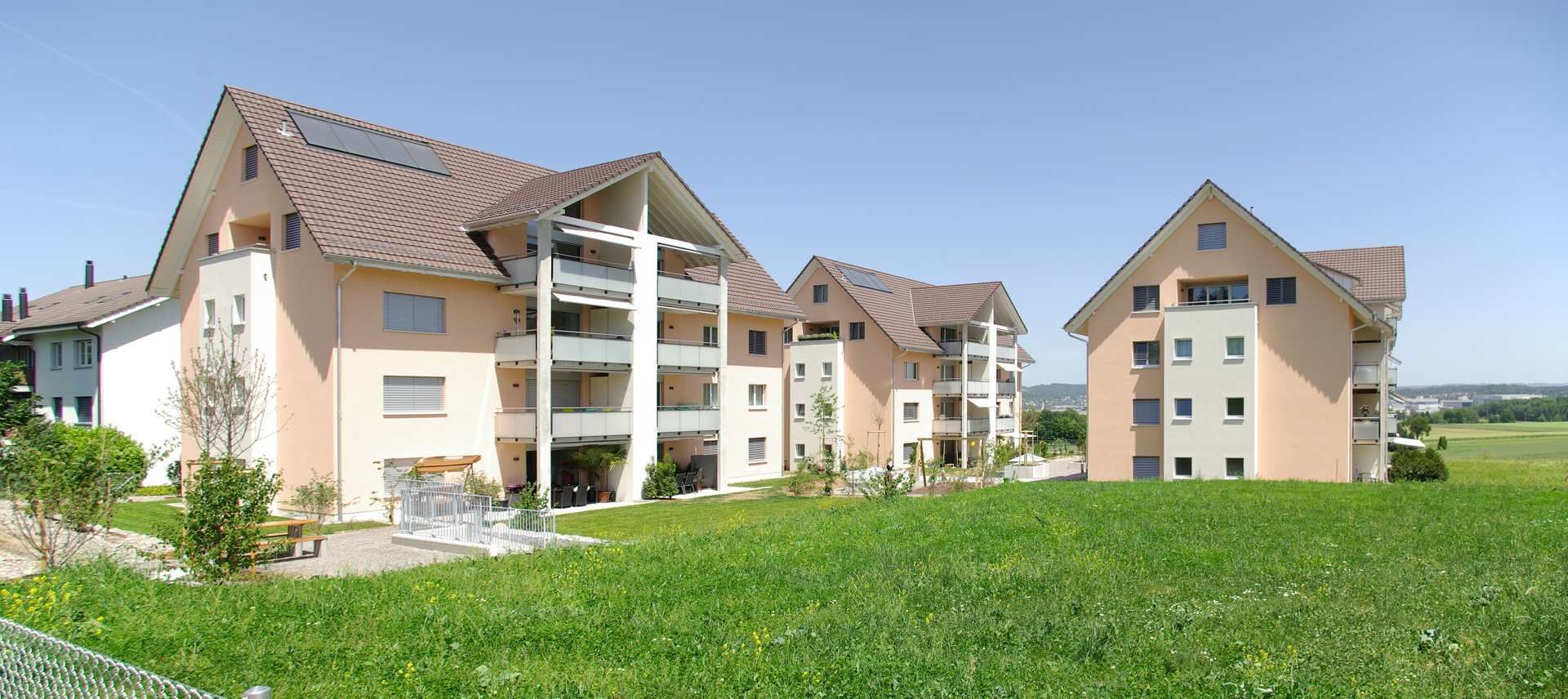 Wohnhäuser mit braunen Dächern, Balkonen und hellbraunen Wänden stehen inmitten einer grünen Wiese.