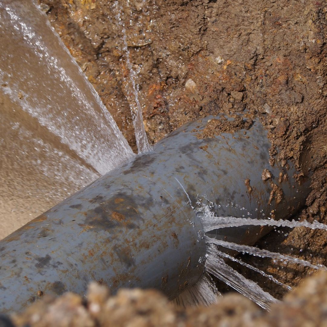Un tuyau gris cassé, d'où jaillit de l'eau, se trouve dans une tranchée de terre.