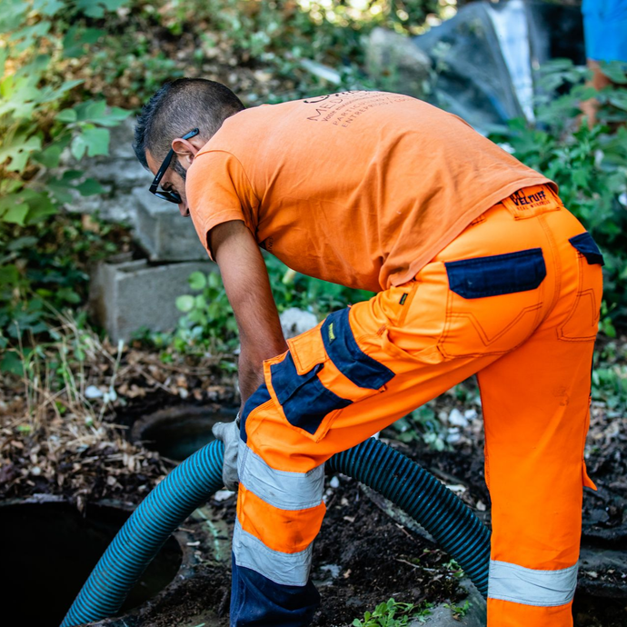 Personne en tenue de travail orange raccordant un tuyau, penchée au-dessus d'une ouverture, à l'extérieur.