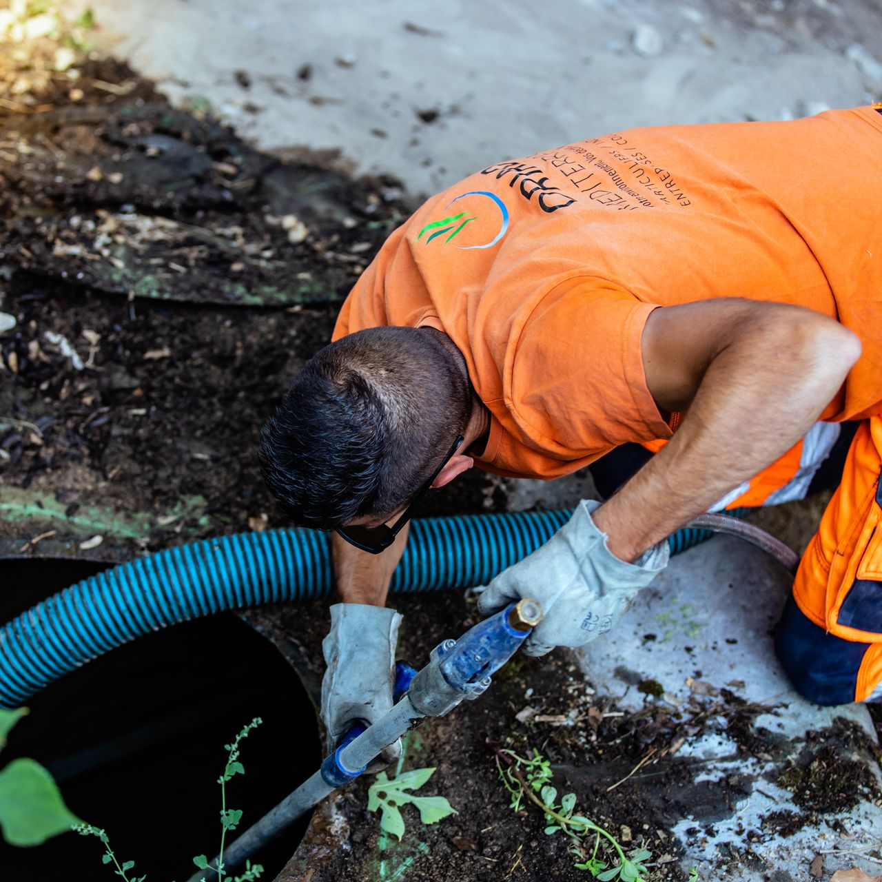 Une personne en chemise orange et gants travaille sur une ouverture de tuyau. Tuyau bleu déployé, à l'extérieur.