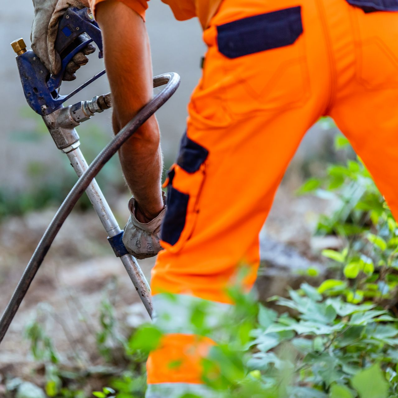 Un ouvrier portant un pantalon et des gants orange utilise un pulvérisateur à l'extérieur.