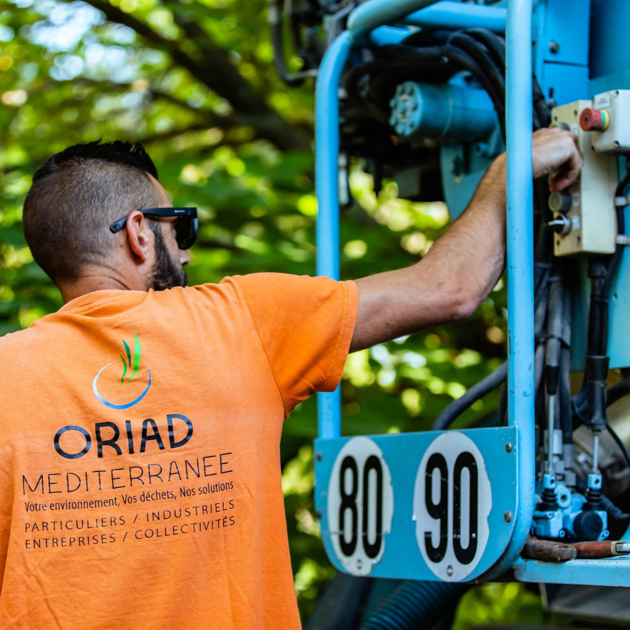 Un homme en tenue orange travaille sur une machine bleue, à l'extérieur.