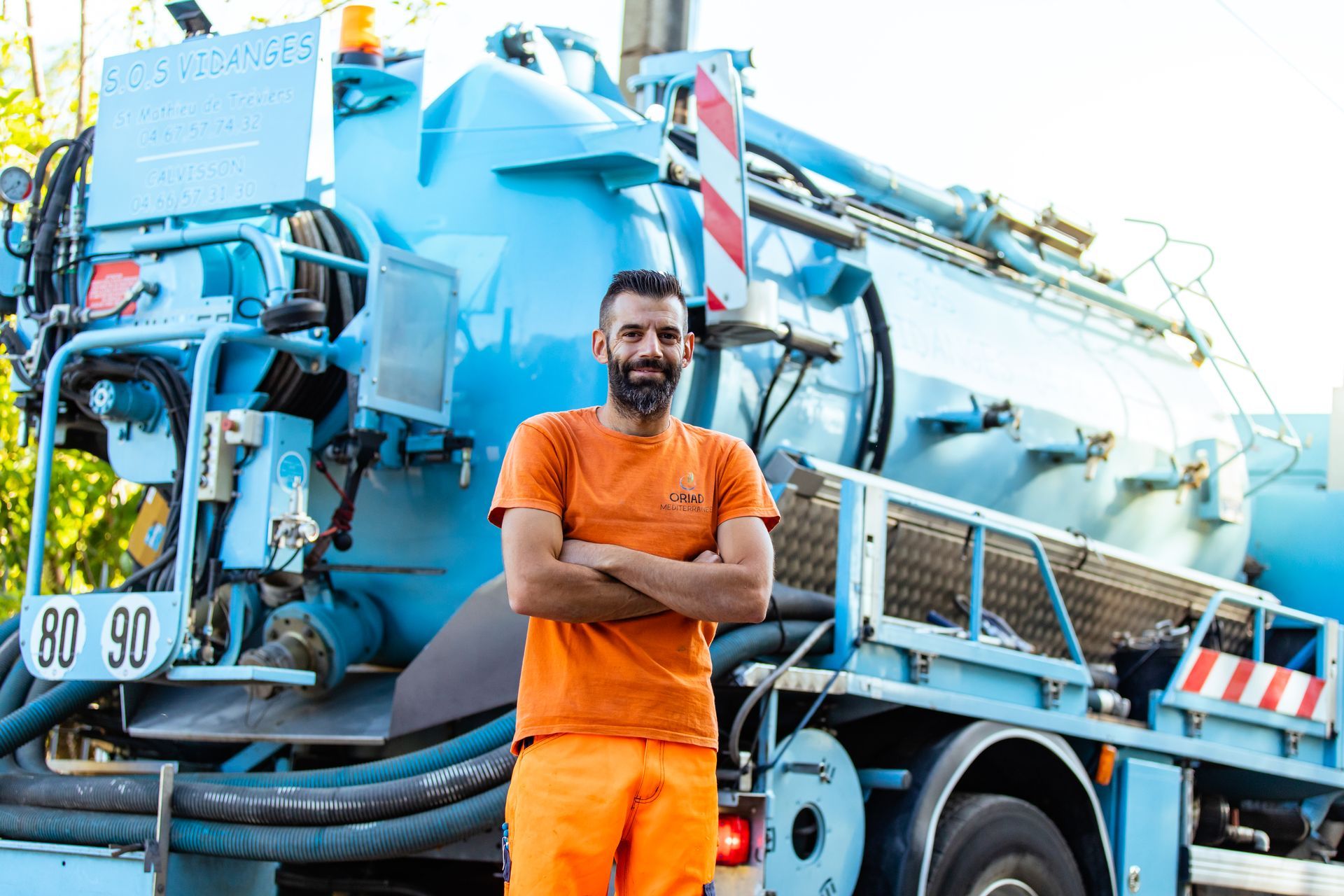 Un homme en tenue de travail orange se tient devant un camion bleu, les bras croisés, en souriant.