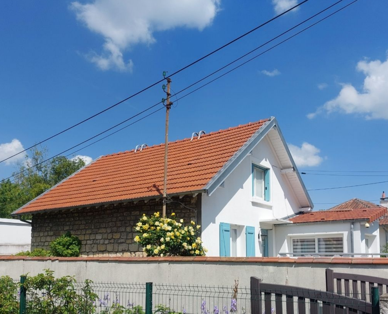 Maison blanche au toit de tuiles orange, mur en pierre, volets bleus et lignes électriques se détachant sur un ciel bleu.